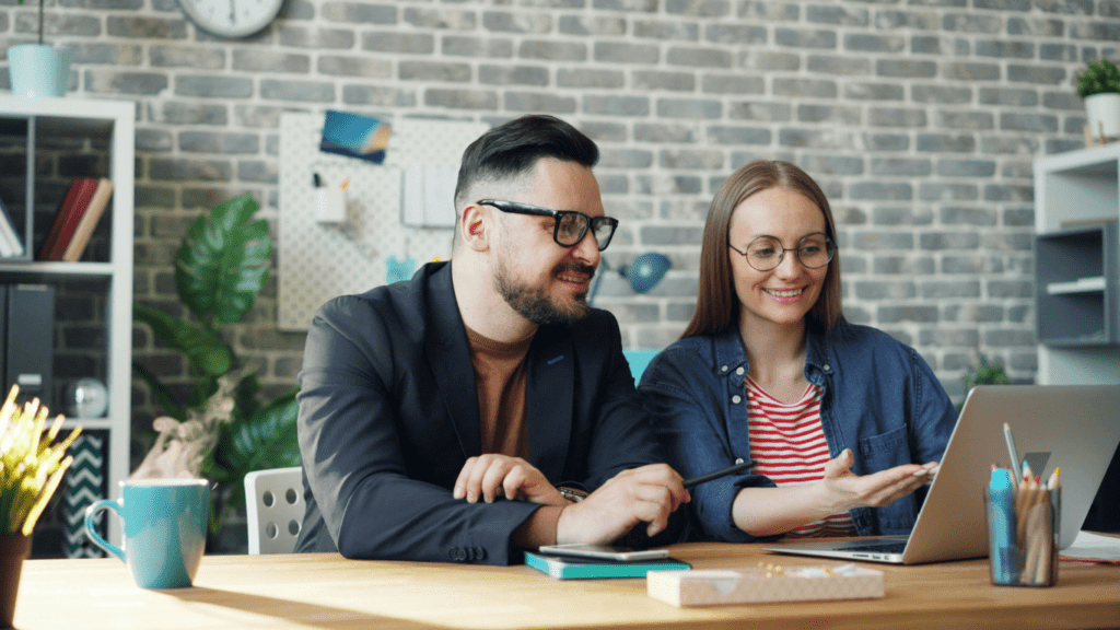 two people sitting at a table with a laptop and papers